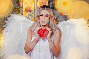 Angel holds a red heart in his hands. Portrait of a girl in a white dress with white wings behind her back
