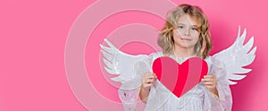 Angel kid with heart. Valentine's day. Blonde cute child with angel wings on a pink studio background. Banner header