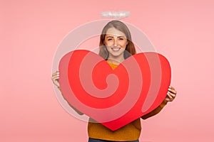 Angel on Valentine`s day. Portrait of lovely ginger girl with halo over head smiling at camera and holding big red heart, symbol