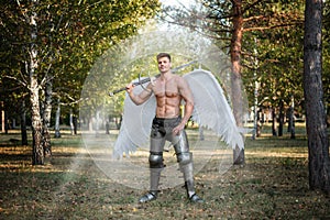 Angel warrior, a bodybuilder in plate armor on his legs with wings behind his back, stands in the beam of light