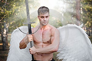 Angel warrior, a bodybuilder in plate armor on his legs with wings behind his back, stands in the beam of light