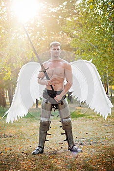 Angel warrior, a bodybuilder in plate armor on his legs with wings behind his back, stands in the beam of light