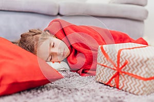 Angelic daughter is sleeping on the carpet, covered with red blanket, with Christmas gift on her side.