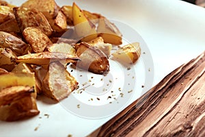 Angle part of white paper and cutting board with delicious baked golden potatoes slices with seasoning from sideways close up. On