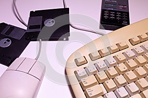 Angle top view of an old computer keyboard, a mouse with their wires, some floppy disks and a remote control on a white background