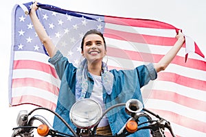 Angle view of beautiful girl sitting on scooter, holding American flag and smiling on sky background
