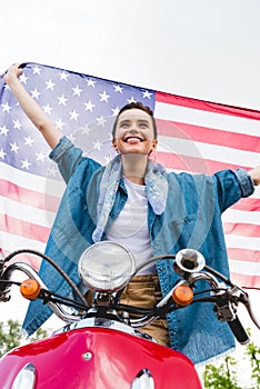 Angle view of beautiful girl standing on red scooter, holding American flag and smiling on sky background