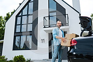 Angle view of cheerful man holding boxes near car and modern house