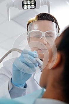 Angle view of dentist in face shield and latex gloves holding dental instrument near african american woman