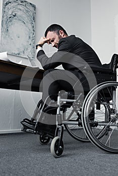 Angle view of depressed disabled businessman sitting in wheelchair at workplace with bowed head