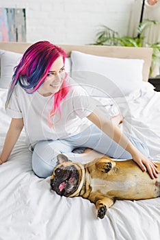 Angle view of girl with colorful hair petting cute french bulldog and looking away while sitting in bed