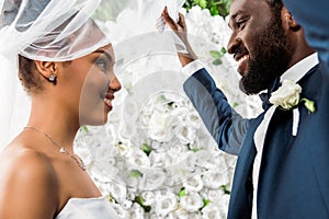 Angle view of happy african american bridegroom touching white veil and smiling near bride and flowers