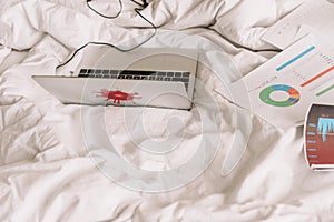 Angle view of laptop and papers with graphs and charts on white bedding