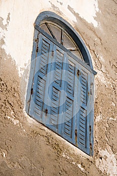 Angled faded blue shuttered window with fanlight set in a peeling plaster wall - dutch tilt