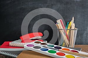 Angled view over a wooden desk with school supplies on it. Stepler, colored pencils, notebooks, calculator, scissors, tapes, paint