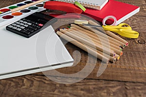 Angled view over a wooden desk with school supplies on it. Stepler, colored pencils, notebooks, calculator, scissors, tapes, paint