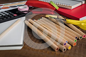 Angled view over a wooden desk with school supplies on it. Stepler, colored pencils, notebooks, calculator, scissors, tapes, paint