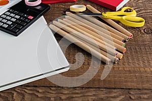 Angled view over a wooden desk with school supplies on it. Stepler, colored pencils, notebooks, calculator, scissors, tapes, paint