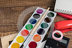 Angled view over a wooden desk with school supplies on it. Stepler, colored pencils, notebooks, calculator, tapes, paints. Back to