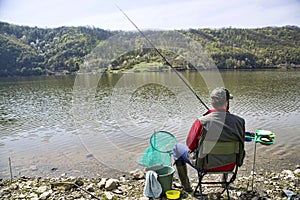 Angler With His Back Towards Camera Sitting On The River Coast Enjoying Angling And Beautiful Nature