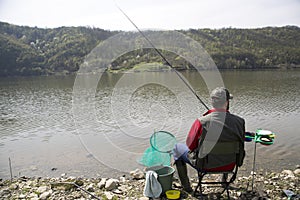 Angler With His Back Towards Camera Sitting On The River Coast Enjoying Angling And Beautiful Nature