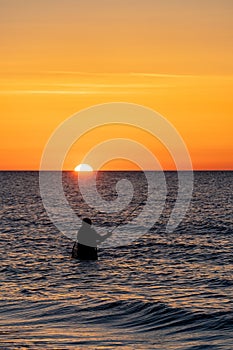 An angler in waterproof trousers fishes in front of a gorgeous orange sunset in the Baltic Sea.