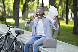 Angry African American man with documents shouting into cellphone near his bike at park