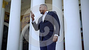 Angry African American young businessman yelling on the phone standing outdoors. Portrait of stressed man in suit