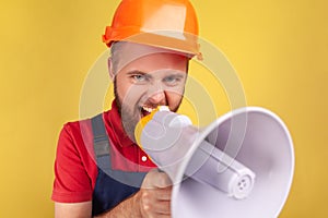 Angry aggressive bearded worker wearing protective helmet and blue overalls holding megaphone.