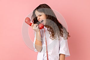 Angry aggressive beautiful little girl shouting into retro phone handset, irritated by call.