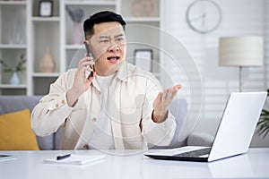 Angry angry and upset man talking on phone at home, asian working remotely from home office sitting at table in living