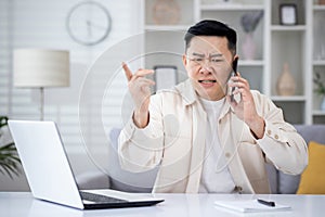 Angry angry and upset man talking on phone at home, asian working remotely from home office sitting at table in living