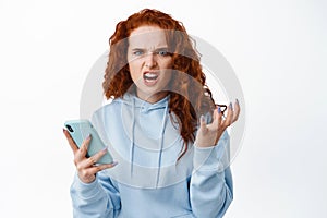 Angry and annoyed redhead girl holding smartphone, frowning and grimacing mad, complaining at something bothering, being