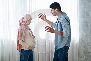 Angry arab man shouting at his pregnant muslim wife, standing near windiw at home, side view