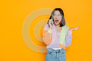 Angry asian girl talking on mobile phone and spreading hand over yellow wall
