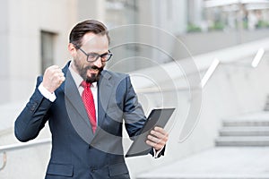 Angry bearded businessman dressed in blue suit threatening with fist to tablet during video conference out of office.