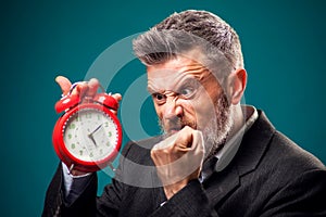 Angry bearded businessman holding alarm clock, shouting and showing fist at it. Time management concept