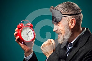 Angry bearded businessman with sleep mask on head holding alarm clock, shouting and showing fist at it. Time management concept