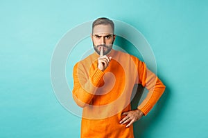 Angry bearded guy telling be quiet, hushing and frowning displeased, shut up gesture, standing over turquoise background