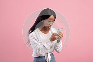 Angry black woman looking at her smartphone with disgust, reading annoying message or news on pink studio background