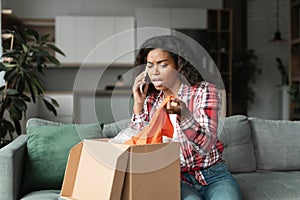 Angry black young woman in casual unpacks parcel, calls by smartphone, swears with store in living room interior