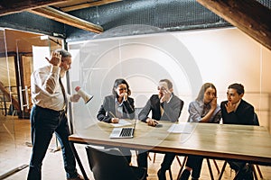 Angry boss with megaphone screaming at employees in office, scared and annoyed colleagues listening at the table