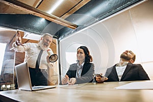 Angry boss with megaphone screaming at employees in office, scared and annoyed colleagues listening at the table