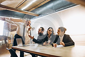 Angry boss with megaphone screaming at employees in office, scared and annoyed colleagues listening at the table