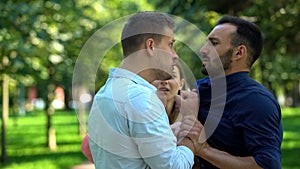 Angry boyfriend pushing man fighting outdoors, girlfriend trying to calm down