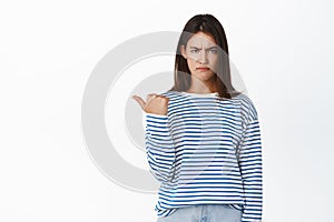 Angry brunette girl pointing left, looking with disapproval and upset pouting face expression, standing in sweater over