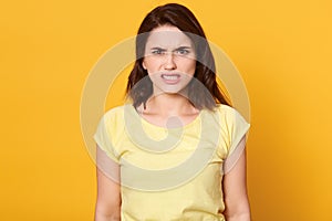 Angry brunette woman looking directly at camera, being very upset, wearing yellow t shirt, standing against bright background,