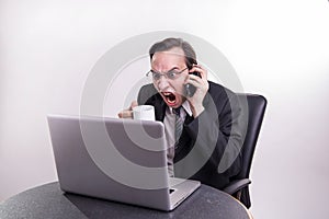 Angry business man talking on a cell phone and screaming on his laptop in the office