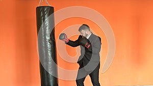 Angry businessman in black suit relieves stress in fitness club. Manager or boss in boxing gloves beats punching bag in