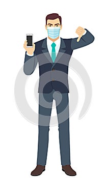 Angry Businessman with medical mask holding a mobile phone and showing thumb down gesture as rejection symbol. Full length